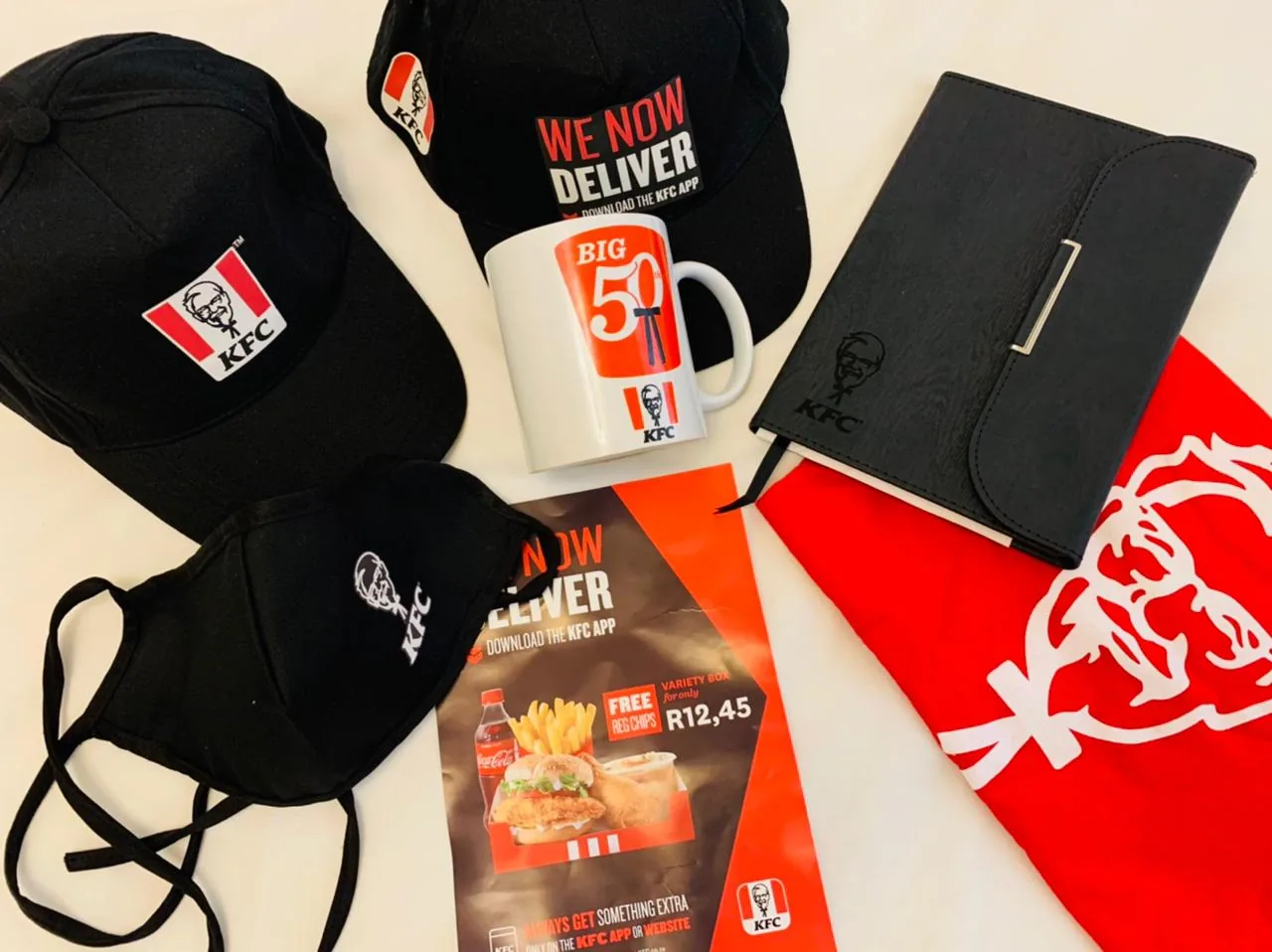 A KFC campaign kit produced by Ithuba Nala: branded caps, a commemorative mug, an embossed notebook, printed face masks, point-of-sale material and a branded bag.